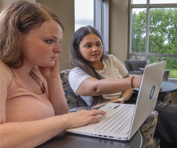 2 students sitting in the library collaborating working on a laptop.