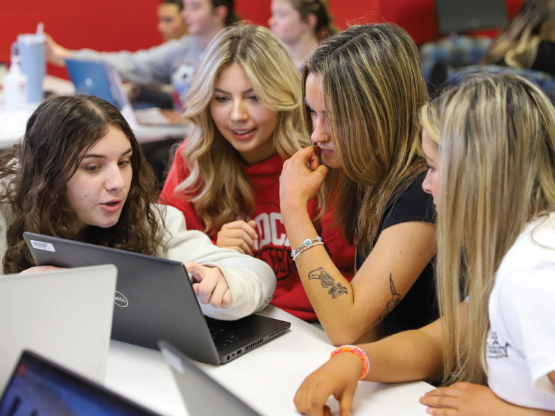 A group of Early College Academy students sitting at a table looking at a laptop working on a project.
