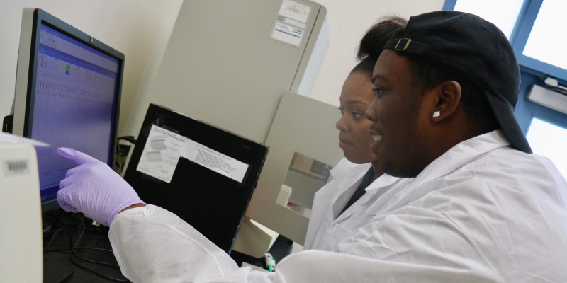 2 students in white lab coats looking at data on the computer