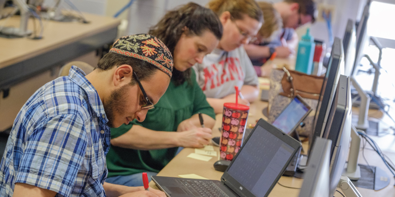Students in a computer classroom sitting in a row writing on post it notes.