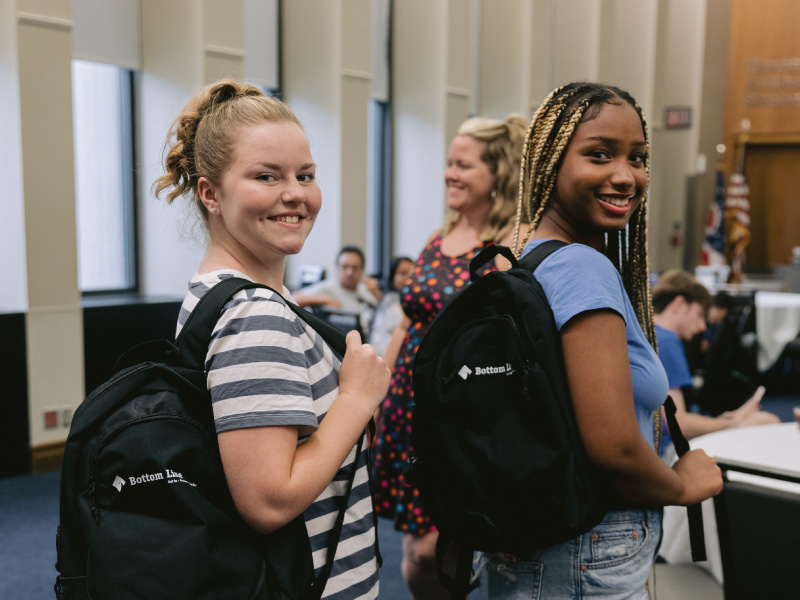 2 students showing off their backpacks with the Bottom Line logo on them.