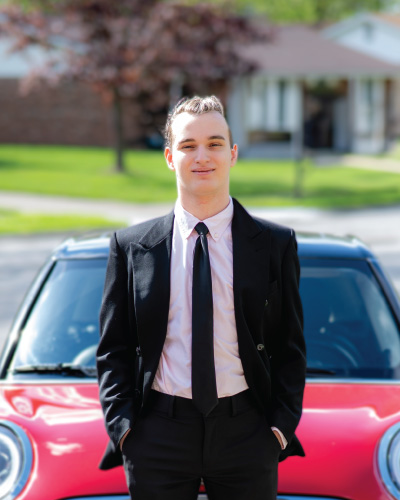 Alex Marcum in a suit and tie standing in front of his red car.